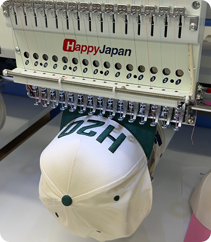 HappyJapan embroidery machine stitching custom lettering onto a white and green baseball cap.