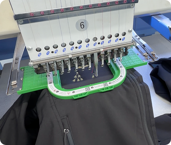Embroidery machine stitching a custom logo onto a black jacket in a professional production workspace.